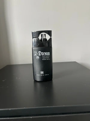 Dream Eau de Toilette svart flaska  - Säljer en Dream Eau de Toilette i en matt svart sprayflaska med vit text och rosdetalj. Flaskan rymmer 100 ml och har en modern, stilren design med praktisk sprayfunktion. Perfekt för dig som gillar unika och snygga parfymflaskor.