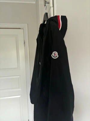 Svart Moncler vindjacka/windbreaker med huva - Svart windbreaker från Moncler med huva och dragkedja. Jackan har Moncler-logga på ärmen och snygga rödvita detaljer på huvan. Perfekt för blåsiga dagar och enkel att matcha med olika outfits.