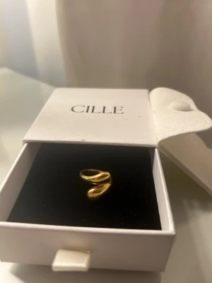 Guldfärgad chunky ring från Cille - Säljer en snygg guldfärgad chunky ring från Cille. Ringen har en modern, öppen design med mjuka, rundade former som ger ett trendigt intryck. Perfekt för dig som gillar statement-smycken och vill addera lite extra stil till din look. Aldrig använd