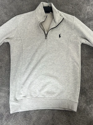 Ljusgrå  tröja Polo Ralph Lauren - Ljusgrå stickad tröja från Polo Ralph Lauren med halv dragkedja och hög krage. Tröjan har en broderad logga i marinblått på bröstet och ribbade muddar vid ärmslut och nederkant. Perfekt för lager på lager och stilren look.