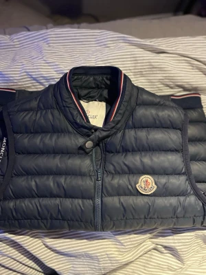 Mörkblå dunväst från Moncler - Snygg mörkblå dunväst från Moncler med klassisk quiltad design och dragkedja framtill. Västen har Moncler-logga på bröstet och ribbad krage med röd, vit och blå rand. Perfekt för lager-på-lager och streetstyle