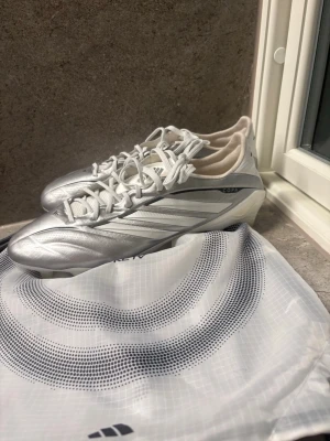 Silvriga Adidas Copa Pure IV fotbollsskor - Snygga silvriga Adidas Copa Pure IV fotbollsskor med vita detaljer och klassisk snörning. Skorna har en modern, strömlinjeformad design och är tillverkade i syntetmaterial för bästa känsla på planen. Perfekta för dig som vill sticka ut på matchen.