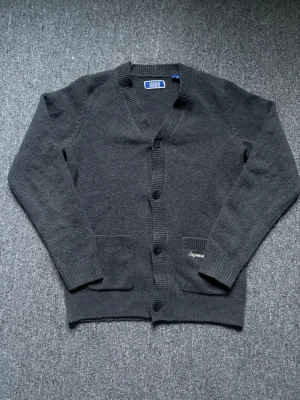 Mörkgrå kofta från Jack & Jones - Mysig mörkgrå kofta från Jack & Jones med broderad text vid fickan. Koftan har v-ringning, knäppning framtill med svarta knappar och två fickor. Ribbstickade muddar vid ärmslut och nederkant ger en snygg finish. Perfekt för lager på lager och chill stil.