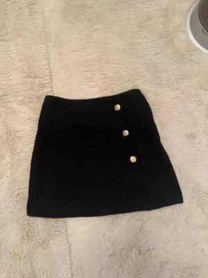 Svart minikjol med guldknappar - Stilren svart minikjol i figurnära modell med tre dekorativa guldknappar framtill. Kjolen har en lätt strukturerad yta och är sydd i ett mjukt tyg som ger en snygg passform. Perfekt att matcha med en crop top eller stickad tröja.
