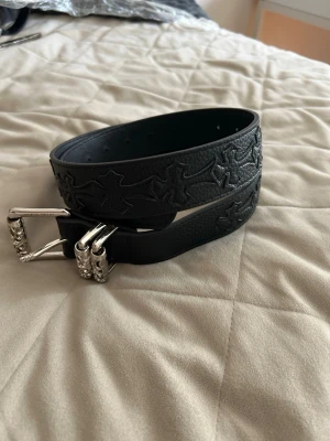 Svart skärp med silverdetaljer - Snyggt svart bälte i skinn med präglade korsmönster längs hela remmen. Spännet är i silverfärgad metall med detaljerade graveringar som ger en unik look. Perfekt accessoar för att lyfta din outfit.