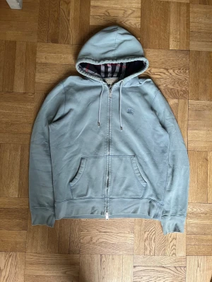 Ljusblå hoodie med dragkedja från burberry - Snygg ljusblå hoodie med dragkedja och huva. Insidan av huvan har ett randigt foder i grått, vitt och rött. Hoodien har två fickor framtill och justerbara snören i huvan. Perfekt för en avslappnad stil.