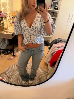 Blommig croppad blus med puffärm - Söt croppad blus med blått blommönster på vit botten. Blusen har v-ringning, knappar framtill och puffiga korta ärmar. Perfekt för vår och sommar när du vill ha en luftig och trendig topp.