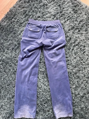 Blå Juicy Couture mjukisbyxor med fickor - Mysiga blå mjukisbyxor med raka ben och bred resår i midjan. Byxorna har snörning framtill och stora fickor både fram och bak, där bakfickorna har lock och knappdetaljer. Perfekta för chill dagar eller när du vill vara extra bekväm.