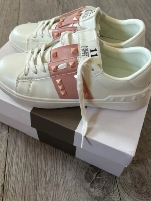 Vita sneakers med rosa nitar - Snygga vita sneakers med breda rosa band över vristen dekorerade med pyramidformade nitar. Skorna har klassisk låg profil, vita snören och en chunky vit sula. Materialet ser ut att vara konstläder och designen är modern och edgy.