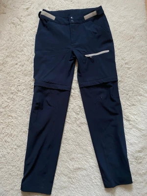 Mörkblå zip-off cargobyxor - Snygga mörkblå cargobyxor med zip-off funktion så du kan göra om dem till shorts. Byxorna har en dragkedjeförsedd ficka på låret och justerbara band i midjan. Perfekta för dig som vill ha både byxor och shorts i ett plagg.