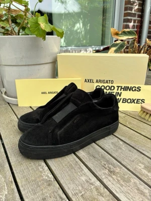 Svarta sneakers från Axel Arigato - Stilrena svarta sneakers från Axel Arigato i mocka med minimalistisk design och ton-i-ton sula. Skorna har låg profil och diskret logga på sidan. Perfekta för dig som gillar en clean och modern look.