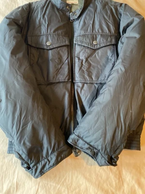 Mörkblå bomberjacka med fickor - Snygg mörkblå vintage bomberjacka med två bröstfickor med lock och knapp, dragkedja framtill och ribbade muddar i nederkant och ärmslut. Jackan har en klassisk bombermodell och är tillverkad i ett glansigt, lätt material som ger en cool look. Jackan har ett litet hål vid armen som inte syns när man går. Priset är inte hugget i sten. 😊 Storleken är XL men passar bäst S,M och L eftersom att den är liten i storlek.