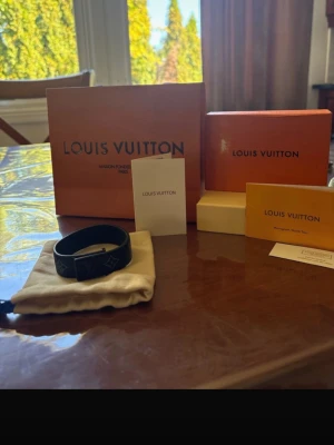 Louis Vuitton svart läderarmband - Snyggt svart armband från Louis Vuitton i präglat läder med klassiska LV-monogramdetaljer. Armbandet har en stilren och modern design med justerbart spänne. Perfekt accessoar för dig som gillar exklusiva märken och vill lyfta din outfit.