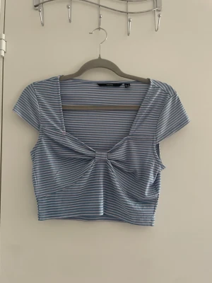 Blåvit randig croppad topp - Snygg croppad topp i blått och vitt med tunna ränder och korta ärmar. Toppen har en twistad detalj framtill och djup v-ringning. Perfekt till jeans eller kjol för en trendig look.