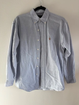 Ljusblå cordskjorta från Polo Ralph Lauren - Ljusblå långärmad skjorta i mjuk manchester från Polo Ralph Lauren. Klassisk button-down krage och broderad logga med polospelare på bröstet. Skjortan har vita knappar och avslappnad passform, perfekt för en clean och stilren look.