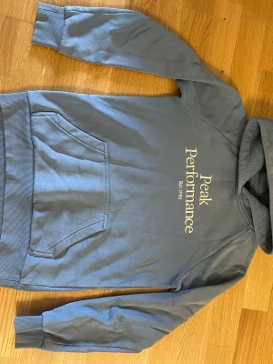 Blå hoodie från Peak Performance - Snygg blå hoodie från Peak Performance med stor logga och text på bröstet. Klassisk känguruficka framtill, ribbade muddar vid ärmar och nederkant samt en skön huva. Perfekt för en avslappnad och sportig stil.