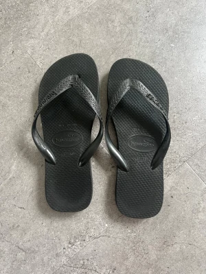 Svarta flip-flops från Havaianas - Klassiska svarta flip-flops från Havaianas med mönstrade remmar och diskret logga på ovansidan. Tillverkade i mjuk och flexibel gummi som ger skön känsla för fötterna. Perfekta för stranden eller sommardagar.