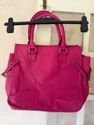 Rosa handväska med axelrem - Snygg knallrosa handväska i konstläder med två handtag och avtagbar axelrem, Åhléns. Väskan har guldfärgade metalldetaljer och en fyrkantig form. Perfekt för dig som vill ha en färgklick till din outfit. Mått 30x33x15cm.