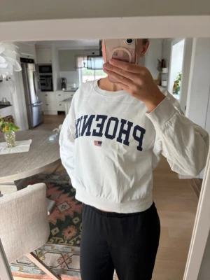 Vit sweatshirt med PHOENIX-tryck - Säljer en vit sweatshirt med mörkblått PHOENIX-tryck och en liten amerikansk flagga på bröstet. Tröjan har rund halsringning, långa ärmar och ribbade muddar vid ärmslut och nederkant. 
