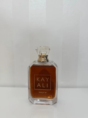Kayali Vanilla 28 parfym - Kayali Vanilla 28 är en lyxig parfym i en elegant, genomskinlig glasflaska med guldiga detaljer och en kristalliknande kork. Doften har en varm, söt vaniljton och flaskan har en modern, kantig form som känns exklusiv och trendig.