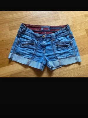 Blå jeansshorts med dragkedjor - Snygga blå jeansshorts med uppvikta benslut och coola dragkedjedetaljer på fickorna. Shortsen har normal passform och klassisk femficksdesign. Perfekta för varma dagar och ger en avslappnad streetstyle-look.
