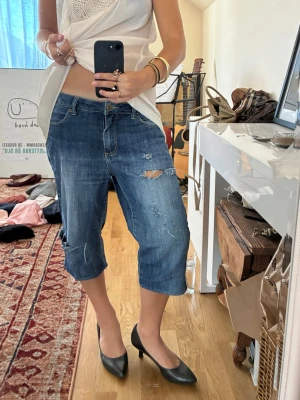Blå caprijeans med slitningar - Säljer ett par blå caprijeans med slitna detaljer och hål på låret. Modellen är loose fit och går ner till under knät. Klassisk femficksdesign och knappgylf. Perfekta för en avslappnad och cool stil. (Egensydda)