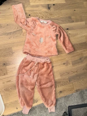 Rosa fluffigt loungewear-set - Supermysigt rosa loungewear-set i mjuk fleece med långärmad tröja och byxor. Tröjan har söta öron på axlarna och ett broderat ansikte framtill. Byxorna har resår och dragsko i midjan samt muddar vid bensluten. Perfekt för chilliga dagar hemma.