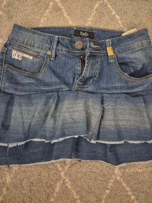 Jeanskjol med volang från D&G - Säljer en blå jeanskjol fejk Dolce & Gabbana med volangdetalj och råa kanter. Kjolen har klassiska fickor fram och bak, samt logotypdetaljer i metall. Perfekt för dig som gillar en edgy och trendig stil.