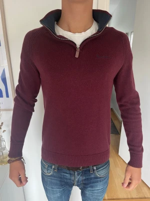 Vinröd halvzip tröja - Snygg vinröd stickad tröja med halvzip och hög krage. Tröjan har ribbade muddar vid ärmslut och nederkant samt diskret broderad logga på bröstet. Model 185 cm, 71kg och bär en S. Hör av er om några frågor