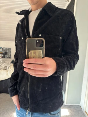 Manchester overshirt  - Säljer en svart overshirt i manchestertyg med dragkedja framtill och två bröstfickor med knappar. Skjortkrage och raka ärmar ger en avslappnad look. Perfekt att slänga över en t-shirt för en cool och enkel stil.