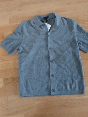 Grå ribbad skjorta med kort ärm - Snygg grå skjorta från H&M med ribbad struktur och korta ärmar. Skjortan har klassisk krage och knäppning framtill med matchande knappar. Perfekt för en avslappnad men stilren look. Materialet känns mjukt och bekvämt.