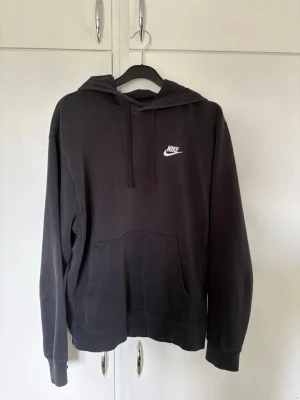 Svart hoodie från Nike - Säljer en svart hoodie från Nike med vit broderad logga på bröstet. Hoodien har huva med dragsko och en stor magficka framtill. Tillverkad i mjukt bomullsmaterial och har ribbade muddar vid ärmslut och nederkant. Perfekt för en avslappnad och sportig stil. Bra skick