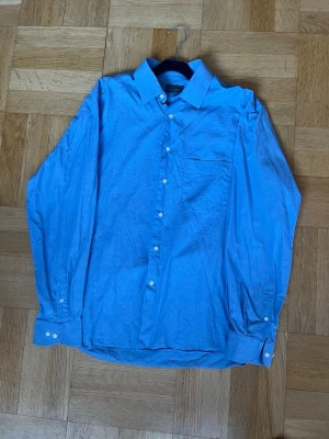 Blå skjorta från Massimo Dutti - Klassisk blå skjorta från Massimo Dutti i 100% bomull. Skjortan har lång ärm, bröstficka och knappar framtill. Perfekt till både jeans och chinos för en clean look.
