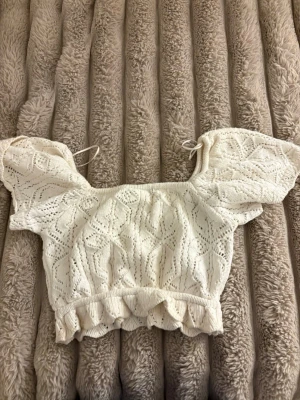 Vit stickad croppad topp Gina Tricot - Supersöt vit stickad croppad topp från Gina Tricot med volangkant och korta puffärmar. Toppen har ett fint hålmönster och knytband vid axlarna. Perfekt för en trendig och somrig look!