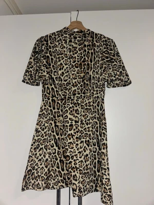 Kort leopardmönstrad klänning - Säljer en kort klänning med leopardmönster i beige, brunt och svart. Klänningen har korta ärmar och rund halsringning. Perfekt för dig som vill sticka ut med ett djuriskt print. Materialet känns mjukt och följsamt, troligen polyester.