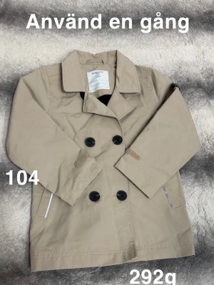 Beige skaljacka för barn - Stilren beige skaljacka för barn med knäppning och klassisk krage. Jackan har knappar, två fickor framtill och detaljer i samma färg som tyget. Perfekt för dig som gillar tidlös och enkel stil.🌸 Använd en gång. Storlek 104