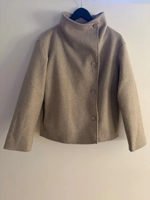 Beige ullrock med hög krage - Stilren beige rock i ullmix med rak passform och hög krage. Rocken har fyra stora knappar framtill och långa ärmar. Enkel design som ger en clean look och passar perfekt till lager på lager under kyliga dagar.