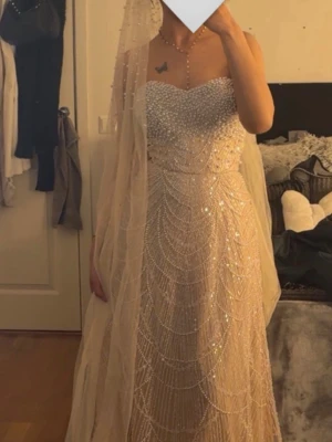 Champagnefärgad långklänning med pärlor - Elegant långklänning i champagnefärg med glittriga pärldetaljer och vackert mönster. Klänningen är axelbandslös och har en svepande kjol med släp. Perfekt för bal eller festliga tillfällen.