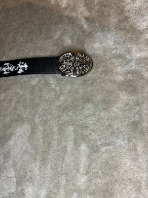 Chrome Svart bälte med vita korsdetaljer - Snyggt chrome svart bälte med vita korsmönster längs hela bandet och ett dekorativt, silverfärgat spänne med detaljerad design. Perfekt accessoar för att lyfta din outfit med en edgy touch. Vet ej om real fick i present