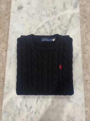 Svart kabelstickad tröja Polo Ralph Lauren - Kabelstickad svart tröja från Polo Ralph Lauren med rund halsringning och röd broderad logga på bröstet. Tröjan har ribbade muddar vid ärmslut och nederkant. Perfekt för dig som gillar klassisk och stilren look.