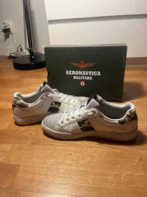 Vita sneakers Aeronautica Militare - Snygga vita sneakers från Aeronautica Militare med kamouflagedetaljer på sidan och hälen. Skorna har lågt skaft, vita snören och en slitstark gummisula. Materialet är en mix av läder och textil för en cool och bekväm look. Använda 2 gånger