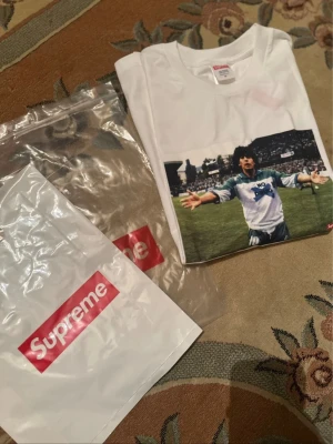 Supreme Maradona - Supreme t-shirt i vitt med ikoniskt Maradona-fototryck på bröstet. Klassisk passform och rund hals. Tillverkad i mjuk bomull, perfekt för dig som gillar streetwear och sportinspirerad stil.