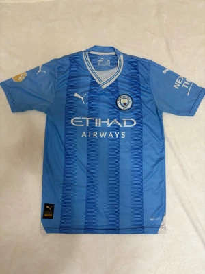 Manchester City  - Snygg ljusblå Manchester City matchtröja från Puma med De Bruyne och nummer 17 på ryggen. Tröjan har klubbmärke, sponsorloggor och vita detaljer. Tillverkad i funktionsmaterial med DryCell-teknologi för bästa komfort under träning eller match.