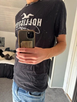 Hollister t-shirt - Säljer en svart t-shirt från Hollister med stort vitt tryck och logga på bröstet. Klassisk passform och rund halsringning. Perfekt till jeans eller shorts för en avslappnad look.