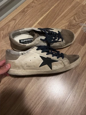 Beige Golden Goose Superstar sneakers - Snygga beige sneakers från Golden Goose med svart stjärna på sidan och mörkblå skosnören. Modellen är Superstar med lågt skaft och slitna detaljer för en cool, avslappnad look. Ovandelen är i läder och mocka, sulan är vit och robust.