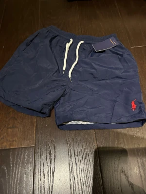 Marinblå badbyxor ralph lauren M - Snygga marinblå badbyxor från Polo Ralph Lauren med vit dragsko i midjan och röd logga broderad på benet. Klassisk modell med innerbyxa i mesh och elastisk midja. Perfekta för stranden eller poolen.