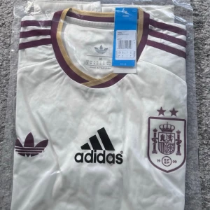 Vit Spanien tröja från Adidas - Säljer en vit Spanien tröja från Adidas med vinröda detaljer och guldkantad krage. Tröjan har Adidas-logga, retro Trefoil-märke och Spaniens landslagsemblem med två stjärnor på bröstet. Perfekt för fotbollsfans som vill sticka ut.