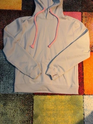 Beige hoodie med rosa snören och tryck - Säljer en beige hoodie med huva och tjocka rosa dragsnören. På huvan finns ett stort graffiti-inspirerat tryck i rosa och grått. Hoodien har en klassisk känguruficka framtill och ribbade muddar vid ärmsluten. Perfekt för en streetwear-look.
