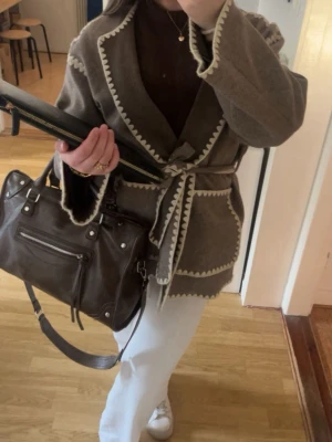 Beige cardiganjacka med knytskärp - Säljer en beige cardiganjacka med dekorativa vita kanter och knytskärp i midjan. Jackan har två stora fickor framtill och är tillverkad i ett mjukt, tjockt material. Perfekt för lager på lager och ger en avslappnad men snygg look.