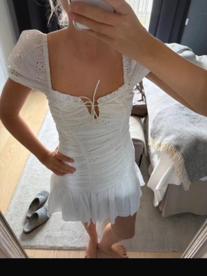 Vit kort klänning med puffärm - Söt vit kort klänning med puffärmar, broderade detaljer och snörning framtill. Klänningen har volangkant nedtill och fyrkantig ringning. Perfekt för sommarens alla tillfällen.